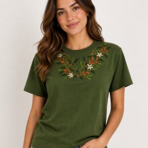 Mujer sonriendo viste una camiseta de color verde oliva con cuello redondo, manga corta y un detallado bordado de flores blancas y hojas de café alrededor del escote.
